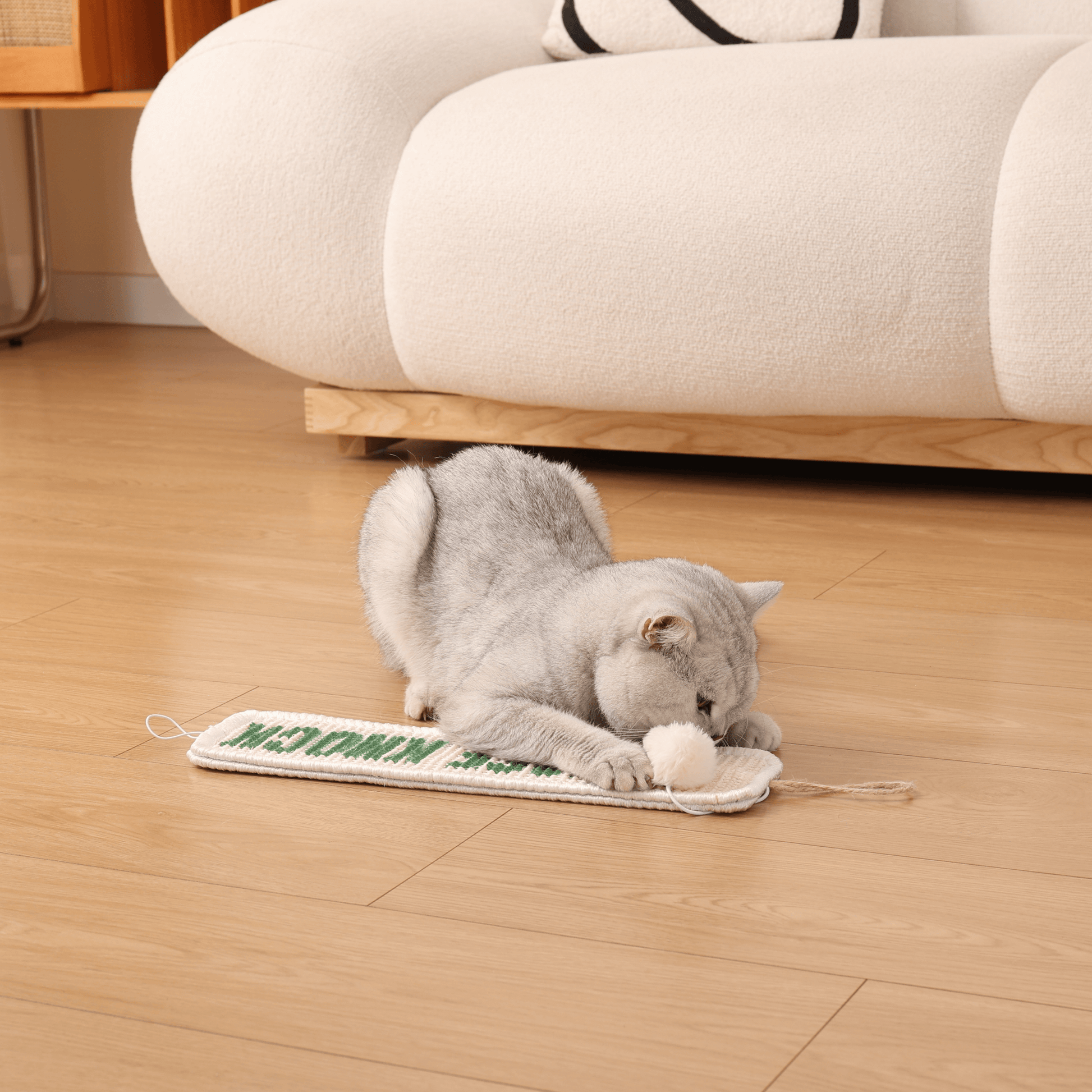 Cat playing with 3-in-1 Hanging Sisal Scratch Mat on wooden floor with a catnip-infused ball, in front of a modern sofa.