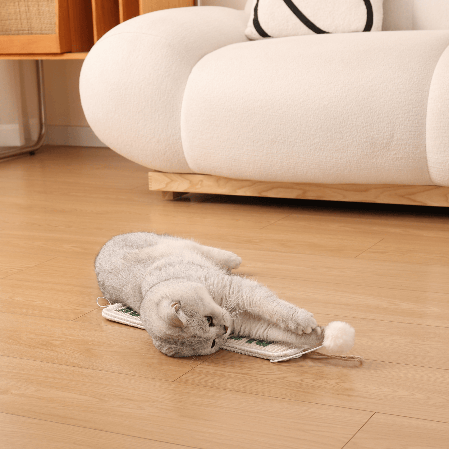 Cat enjoying Furrther's 3-in-1 Hanging Sisal Scratch Mat with catnip ball, lying on a wooden floor by a cozy white sofa.